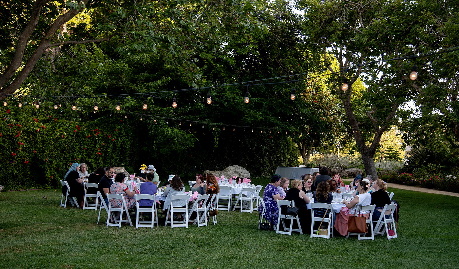 Guests seated at round tables during an outdoor garden dinner under string lights, surrounded by lush greenery.