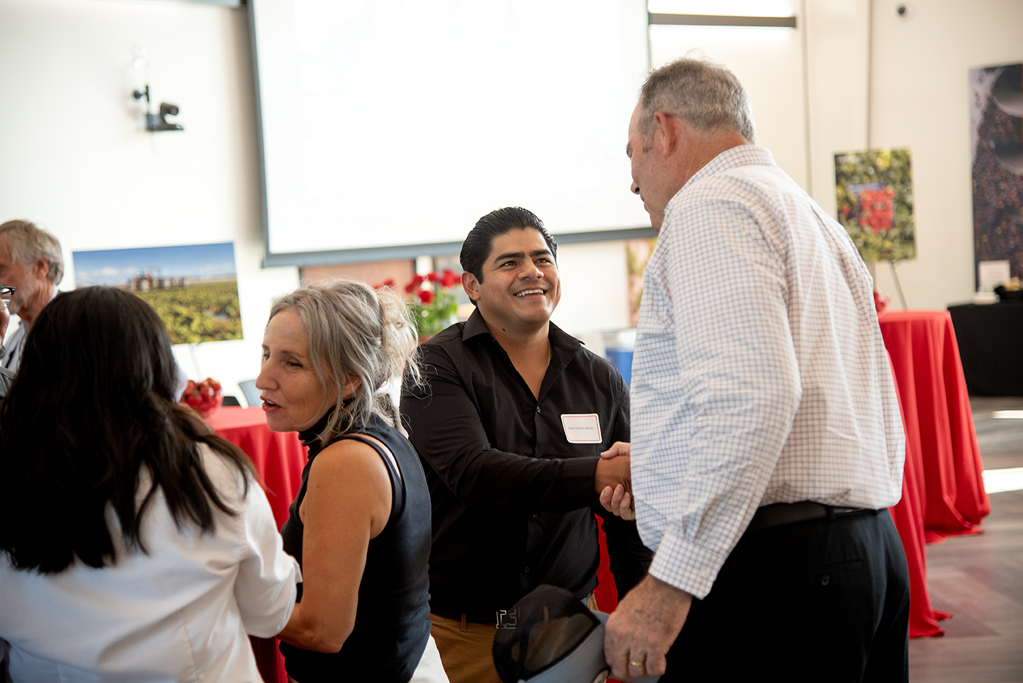 A smiling man shakes hands with another attendee during a networking event in a bright conference room decorated with red tablecloths and agricultural-themed photos.