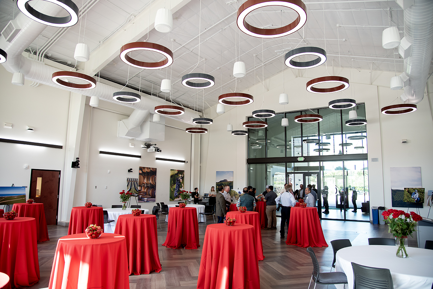 Wide-angle view of a modern event space with circular ring lights on the ceiling, red tablecloth-covered cocktail tables with bowls of strawberries, and guests networking near the glass entrance.