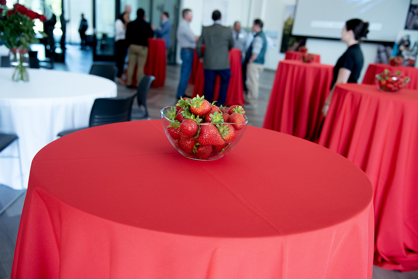 Clear glass bowl filled with fresh strawberries on a red tablecloth, with people mingling in the background at a networking event.