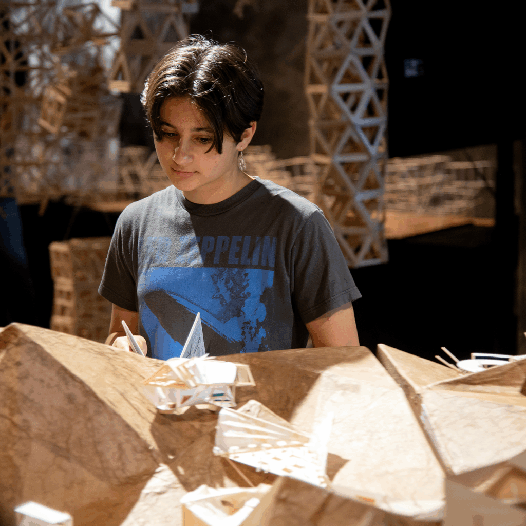 Architecture student at Cal Poly working on a geometric model with wooden structures in the background.