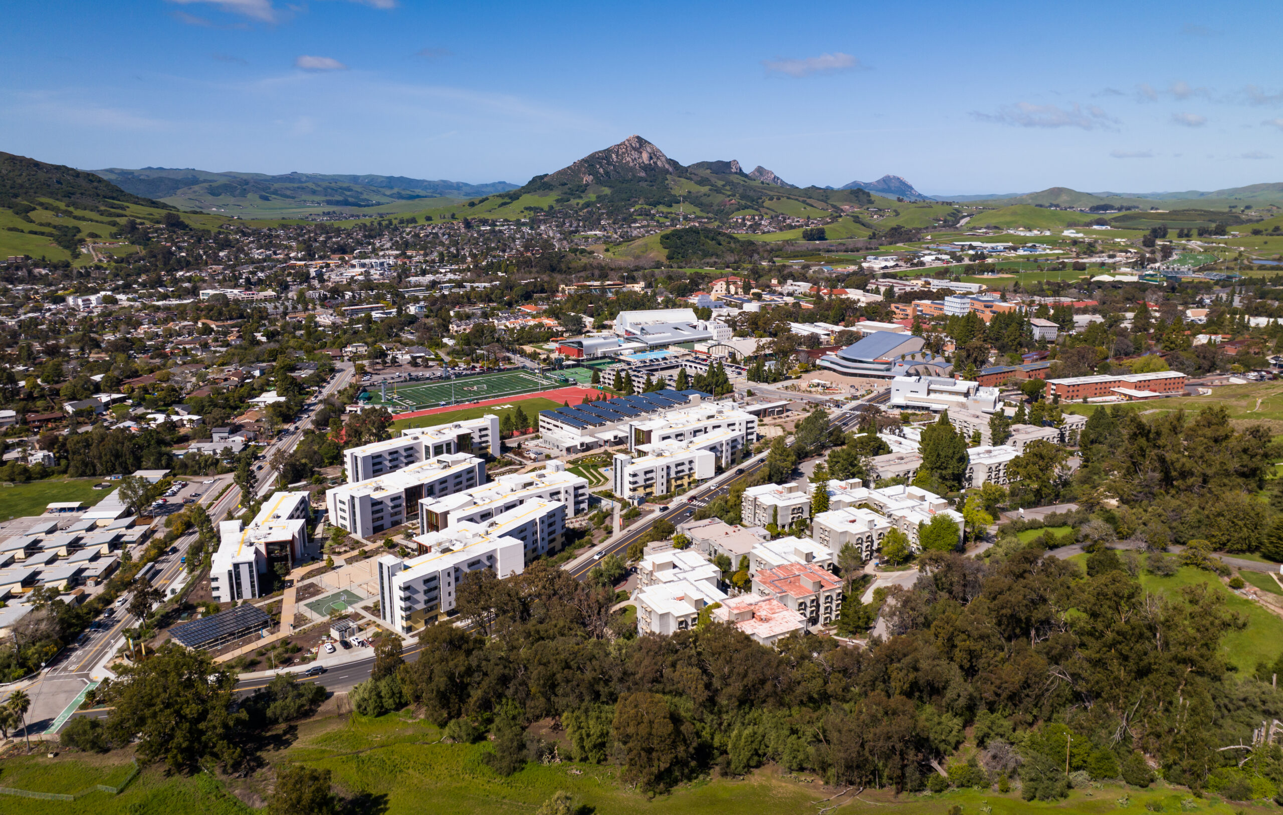 Aerial view of Cal Poly San Luis Obispo campus with dorms, sports fields, and scenic hills under a clear blue sky.