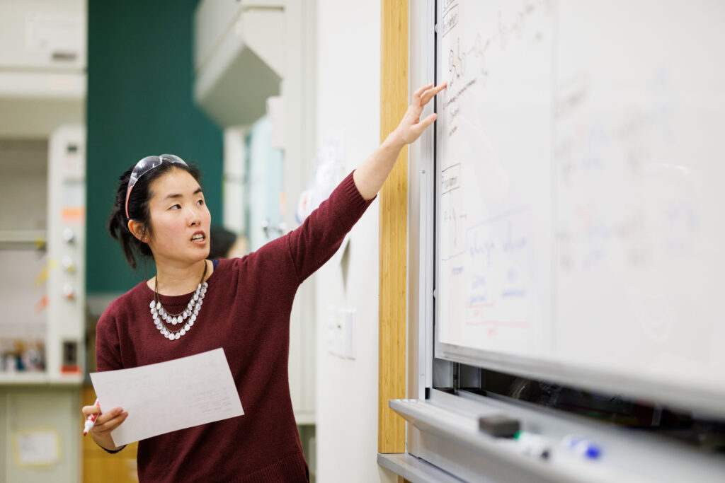 Cal Poly instructor pointing at complex chemistry equations on a whiteboard while holding a marker and notes