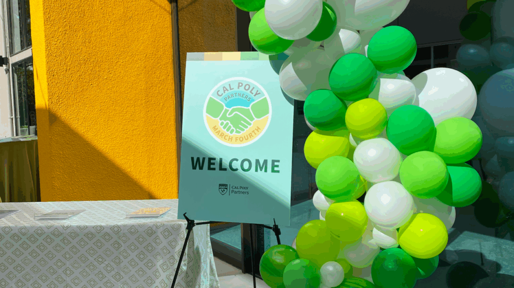 A colorful Cal Poly March Fourth welcome sign stands next to green, yellow, and white balloons inside a bright building lobby.
