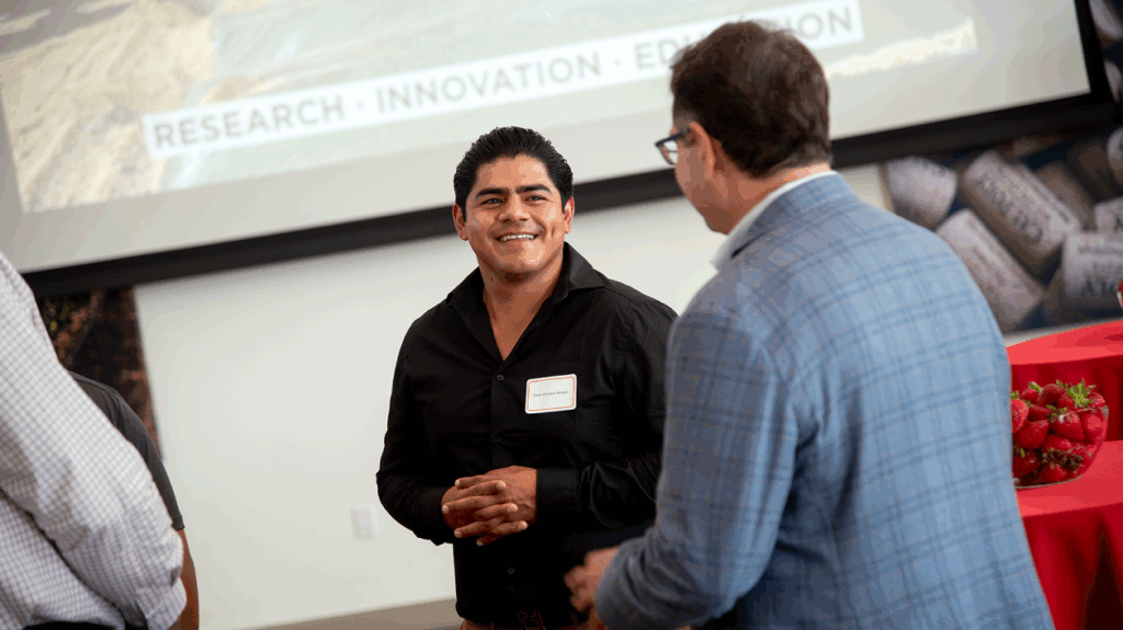 A smiling man in a black shirt speaks with another person during a Cal Poly networking event, with a screen and strawberries in the background.
