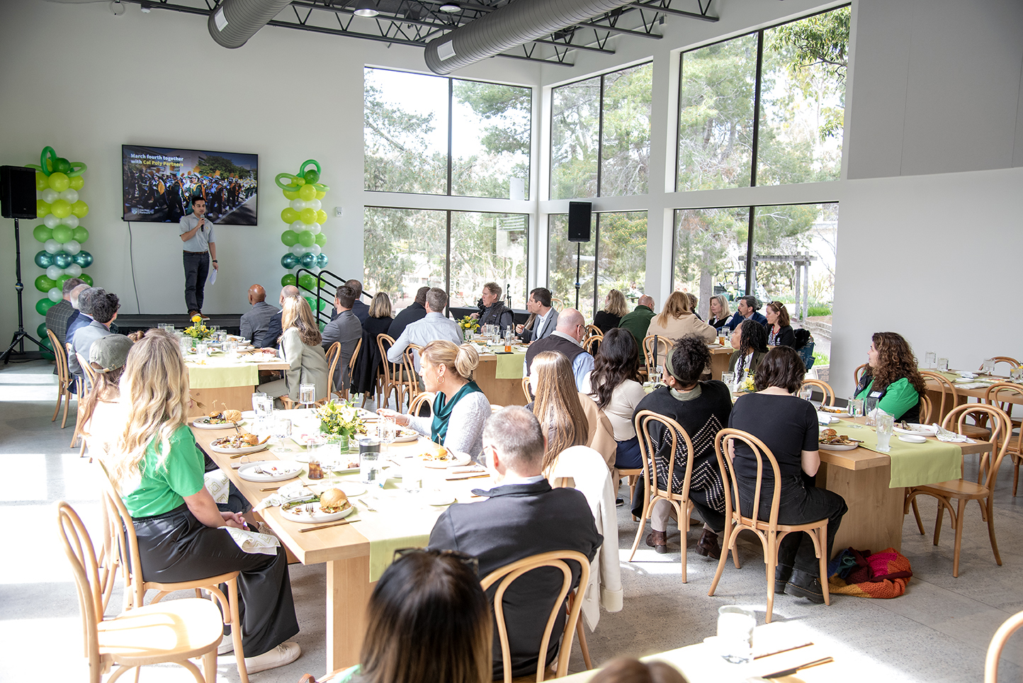 People attending a luncheon event inside the Cal Poly Partners Pavilion with large windows and green balloon decorations.