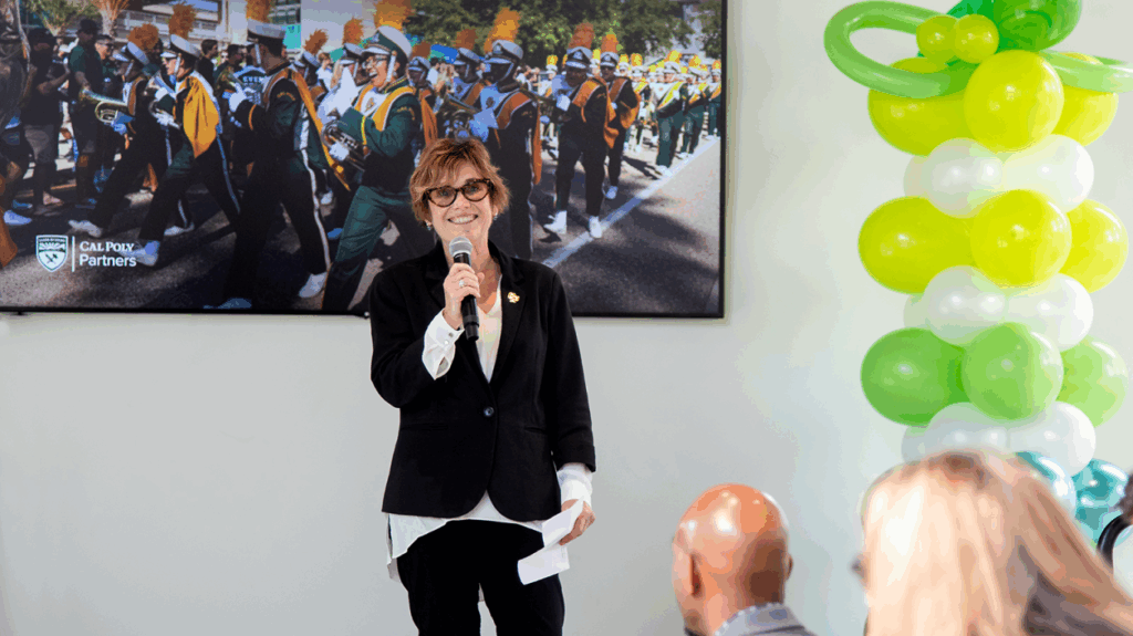Woman speaking at podium with Cal Poly marching band poster behind her.