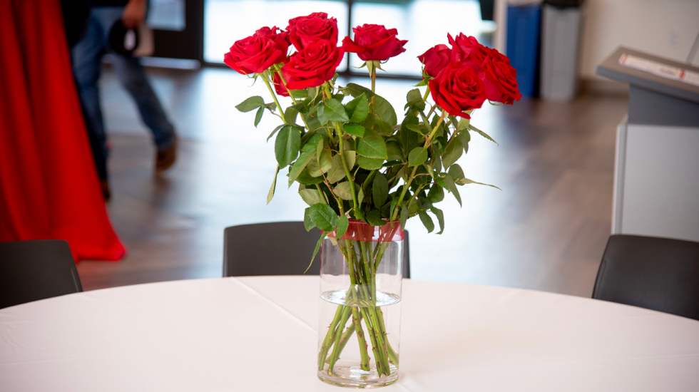 A bouquet of red roses in a clear glass vase sits on a white tablecloth, arranged as a centerpiece in an indoor setting.