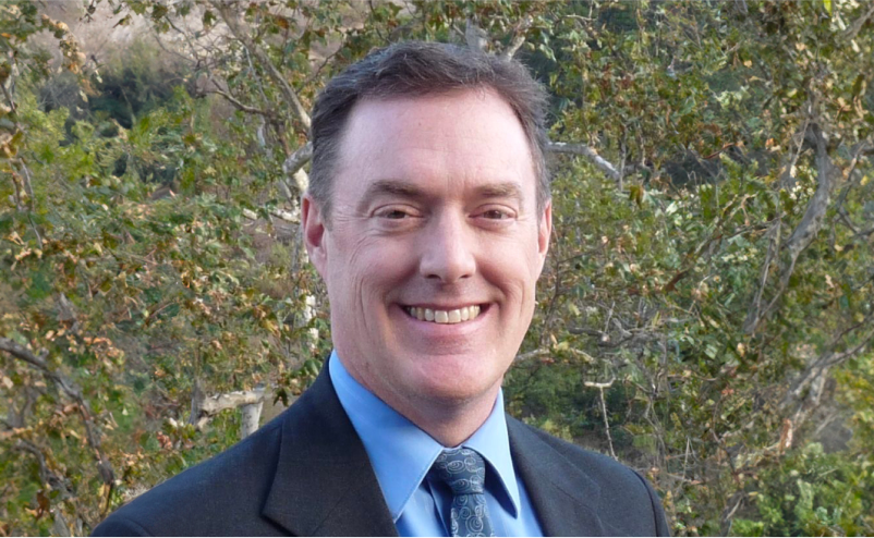 Headshot of Scott in a suit and tie outdoors with trees in background