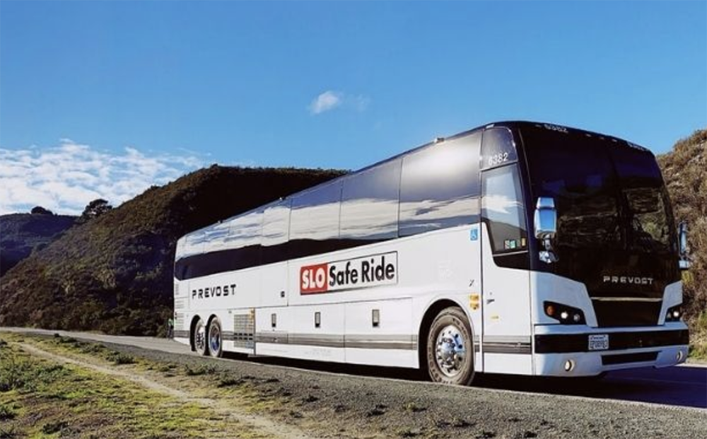 SLO Safe Ride charter bus parked on a roadside with scenic hills and a bright blue sky in the background.