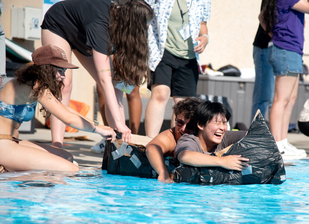 Students laughing and paddling in a duct-taped cardboard boat during a pool competition at Cal Poly
